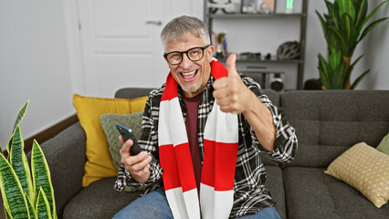 Sticker - Smiling middle-aged man with grey hair at home showing thumbs up while holding a smartphone, conveys positivity and technology usage.