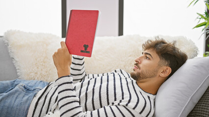 Sticker - Young arab man reading book lying on sofa at home