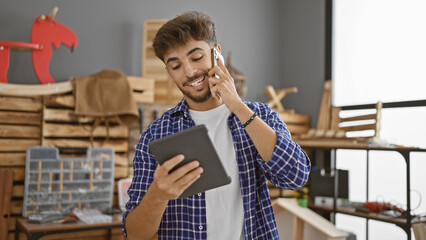 Sticker - Handsome young arab man carpenter talking on smartphone, beaming with smiling face, engrossed in woodworking on touchpad indoors at his carpentry workshop studio