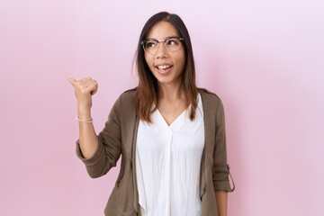 Wall Mural - Middle age chinese woman wearing glasses over pink background smiling with happy face looking and pointing to the side with thumb up.