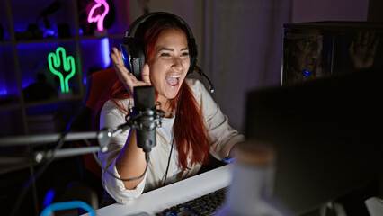 Smiling young redhead woman streamer engaging in a lively video call amidst her invigorating gaming room, immersed in a virtual gaming world, surrounded by the humming of her gear.