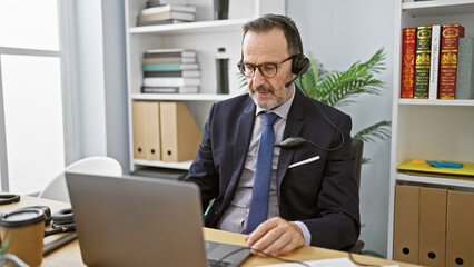 Wall Mural - Hispanic middle-aged man with grey hair, focused on business work in the office, professionally speaking to a customer, providing aid through laptop. concentrated, successful worker using headset