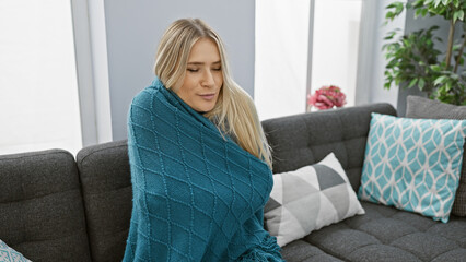 Sticker - Beautiful young blonde woman, freezing in the cozy warmth of her apartment, sitting on the sofa swaddled in a blanket fighting the cold fever of flu