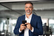 © Stock 4 You - Smiling mature Latin or Indian businessman holding smartphone in office. Middle aged manager using cell phone mobile app, looking at camera. Digital technology application and solutions for business.