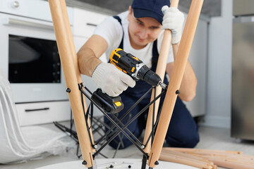 Man in a blue jumpsuit is using an electric drill to assemble a table.