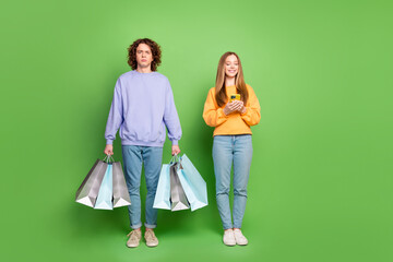 Poster - Full body portrait of cheerful girl use smart phone unsatisfied guy hold store shop bags isolated on green color background