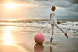 Hairless ballerina with alopecia in white futuristic suit jumps next to pink sphere and soaring along seashore at sunset sea, metaphoric surreal scene exudes confidence, hope and unique beauty