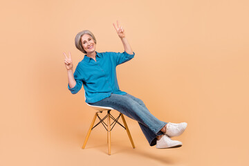 Wall Mural - Full size photo of satisfied relaxed woman dressed stylish shirt sit on chair showing v-sign symbol isolated on pastel color background