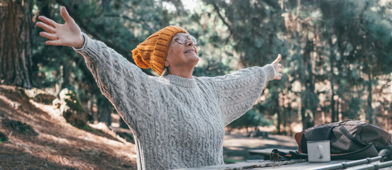 Head shot portrait close up of middle age woman enjoying and relaxing sitting at table in the nature in the forest of mountain. Old female person opening arms and closing eyes feeling free. Freedom 