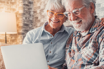 Wall Mural - Cute couple of old people sitting on the sofa using laptop together shopping and surfing the net. Two mature people wearing eyeglasses in the living room enjoying technology. Portrait of seniors laugh