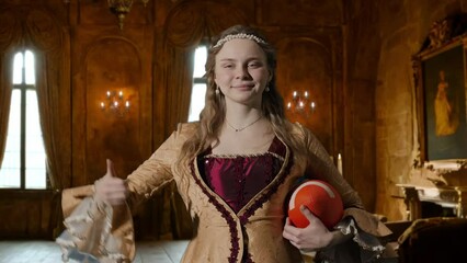 Wall Mural - Woman in ancient outfit on background of historic interior. Female in renaissance style dress holding american football ball smiles shows thumb up.