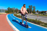 Fototapeta Tulipany - Young latin man riding out of the park on an e-scooter on a blue bike path. Commuting through the town via powered standing scooter. Copy space, background.