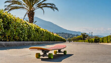 A Skateboard Sits Idly On The Side Of The Road, Waiting For Its Rider. Street View With Palm And Lonely Long Board