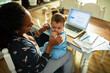 © Marko Geber - Multitasking mother holding baby while working from home