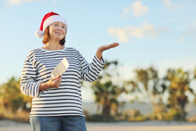 Happy Middle Aged Woman In Red Christmas Santa Claus Hat Holds Passport, Boarding Ticket, Presenting Empty Copy Space On Open Hand Palm On Seashore With Palms. New Year Holidays, Winter Vacation.