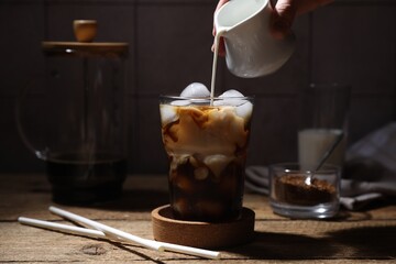 Wall Mural - Woman pouring milk into glass with refreshing iced coffee at wooden table, closeup