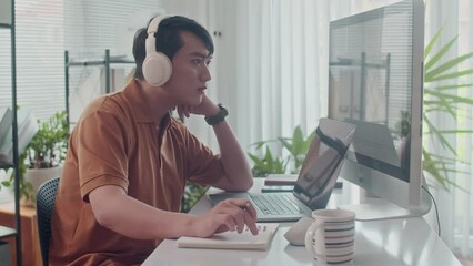 Side view of young Asian tester in headphones working on computer at modern office