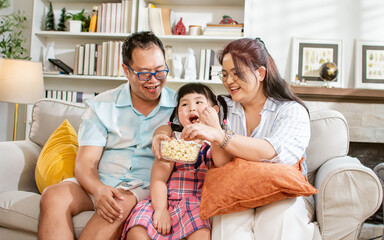 Portrait of three members family, father, mother, daughter, eating popcorn, watching television, movies together in living room at cozy home with happiness, fun. Lifestyle, Education, Kid Concept.