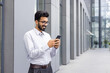 © Liubomir - A young businessman from outside an office building uses the phone, a man smiles and browses social networks, a company employee in a shirt with a beard walks around the city.