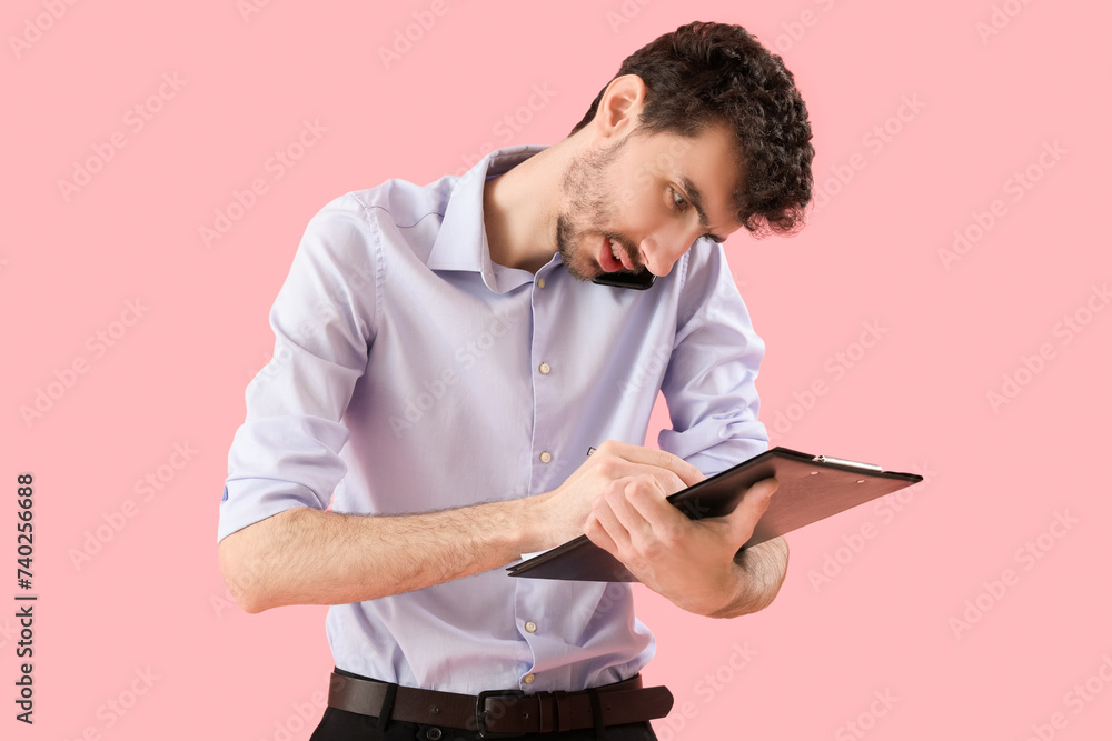 Young businessman with clipboard talking by mobile phone on pink background