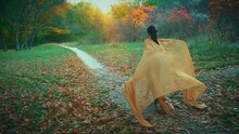 Fantasy Happy Fairy Woman Runs In Autumn Tree Forest. Princess Girl Green Golden Long Vintage Dress Silk Fabric Waving In Slow Motion. Back Rear View Mystery Nymph Along Path. Nature Sun Divine Light