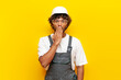 shocked Indian builder in hard hat and overalls covers his mouth with his hand on yellow isolated background, surprised Indian foreman in uniform looks at camera in amazement