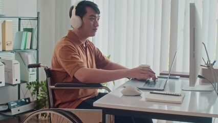 Tilt up portrait of smiling Asian coder in wheelchair turning to camera sitting in front of big monitor at office desk