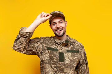 Wall Mural - young Ukrainian army soldier in camouflage pixel uniform smiling on yellow isolated background, Ukrainian military cadet putting on cap