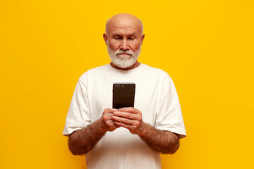 serious old bald grandfather in white t-shirt using smartphone on yellow isolated background