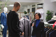 © DC Studio - Fashion boutique arab customer asking asian woman assistant for opinion while choosing formal jacket. Clothing store seller and buyer holding apparel on hanger and discussing style