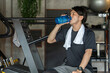 フィットネスジムで休憩・インターバル中に水・プロテインを飲む若いアジア人男性（水分補給）
