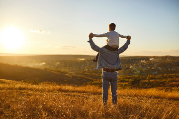 Fathers day. Back view of a little child boy sitting on his fathers shoulders holding hands and looking into the distance enjoying sunset. Father walking with son outdoors.