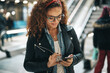 © Flamingo Images - Young woman with curly hair and glasses standing in a metro station reading text messages