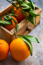 Ripe Oranges With Fresh Green Leaves Attached Are Displayed, Some Resting On A Textured Surface And Others Nestled In A Wooden Crate, Evoking A Sense Of Fresh Harvest