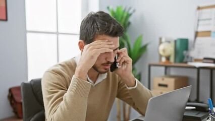 Wall Mural - A focused hispanic man speaks on the phone in a modern office, showcasing professionalism and determination.