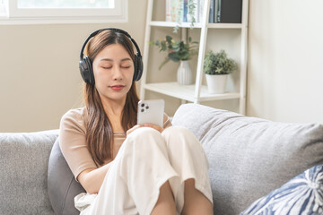 Relaxing asian young woman using smart phone listening to music, enjoy song or watching videos, podcast on mobile with white wireless headphones sitting on sofa, couch at home, chill out and leisure.