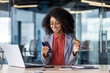 © Liubomir - Satisfied african american female keeping debit card and cellular phone in hands by desk with computer and notebook. Pleased lady entering bank data in application for completing cashless payment