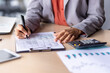 © Liubomir - Close up of unrecognizable black woman in formal outfit filling up tables on clipboard next to electronic calculator. Responsible administrator entering data by hand with pen on wooden desk