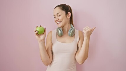 Canvas Print - Happy young woman in sportswear, blue eyes sparkling, rocks out to her headphones while munching an apple, pointing thumb side-ways. all over a sassy pink isolated background.