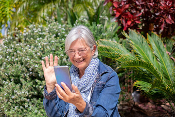 Sticker - Video call concept. Cheerful modern senior white-haired woman with eyeglasses sitting in outdoor garden under the sun using phone in video chat