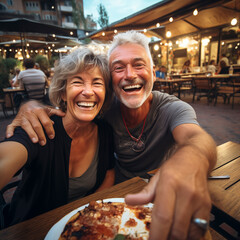 Happy senior old couple have fun eating a pizza together outdoor in traditional italian pizzeria restaurant sitting and talking and laughing. People enjoying food and elderly lifestyle. Tourism