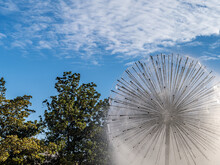 Dandelion Water Fountain In A Park With A Partly Cloudy Blue Sky And Treetops In The Background.