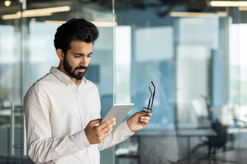 Indian young male programmer standing in the office center indoors and looking focused at the screen of the tablet he is holding in his hands and wearing glasses