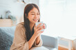 © oatawa - Joyful young asian female enjoying a cup of coffee while sitting on the the sofa at home, Cosy scene, Smiling pretty woman drinking hot tea in winter.