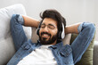 © Prostock-studio - Content indian man wearing wireless headphones reclining peacefully on couch at home