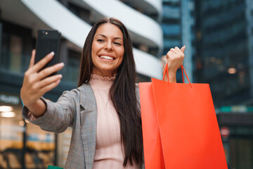 Beautiful smiling young businesswoman sitting in front of modern shopping mall and taking a selfie with items she just bought.