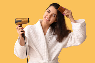 Wall Mural - Beautiful young happy woman in bathrobe with hair dryer and comb on yellow background