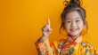 5 year old girl in a fresh and neat cheongsam Smiling brightly as he pointed up and to the right. With a bright yellow backdrop. It adds a delightful touch.