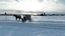 Amish Horse And Buggy Passing On Snow Covered Road In Rural USA Countryside In Winter. Aerial Tracking Shot.