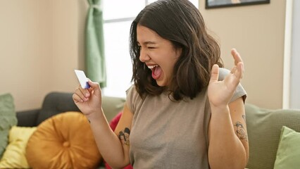 Canvas Print - Victorious young hispanic woman ecstatically celebrating a winning achievement at home! sitting on the sofa, credit card in hand, her beautiful smile speaks for her joy and success.